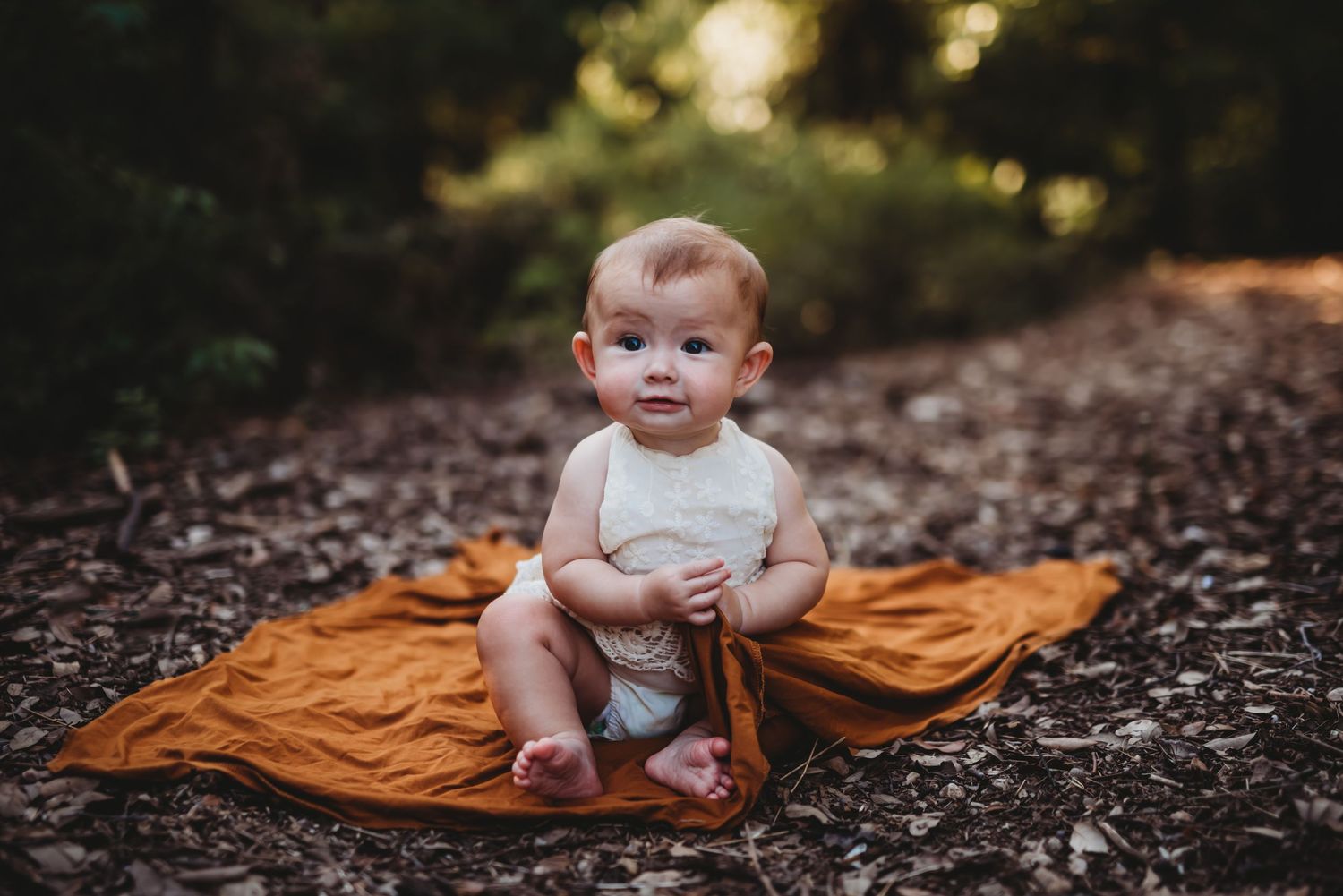 Baby in white dress sitting on orange blanket surrounded by fallen leaves.