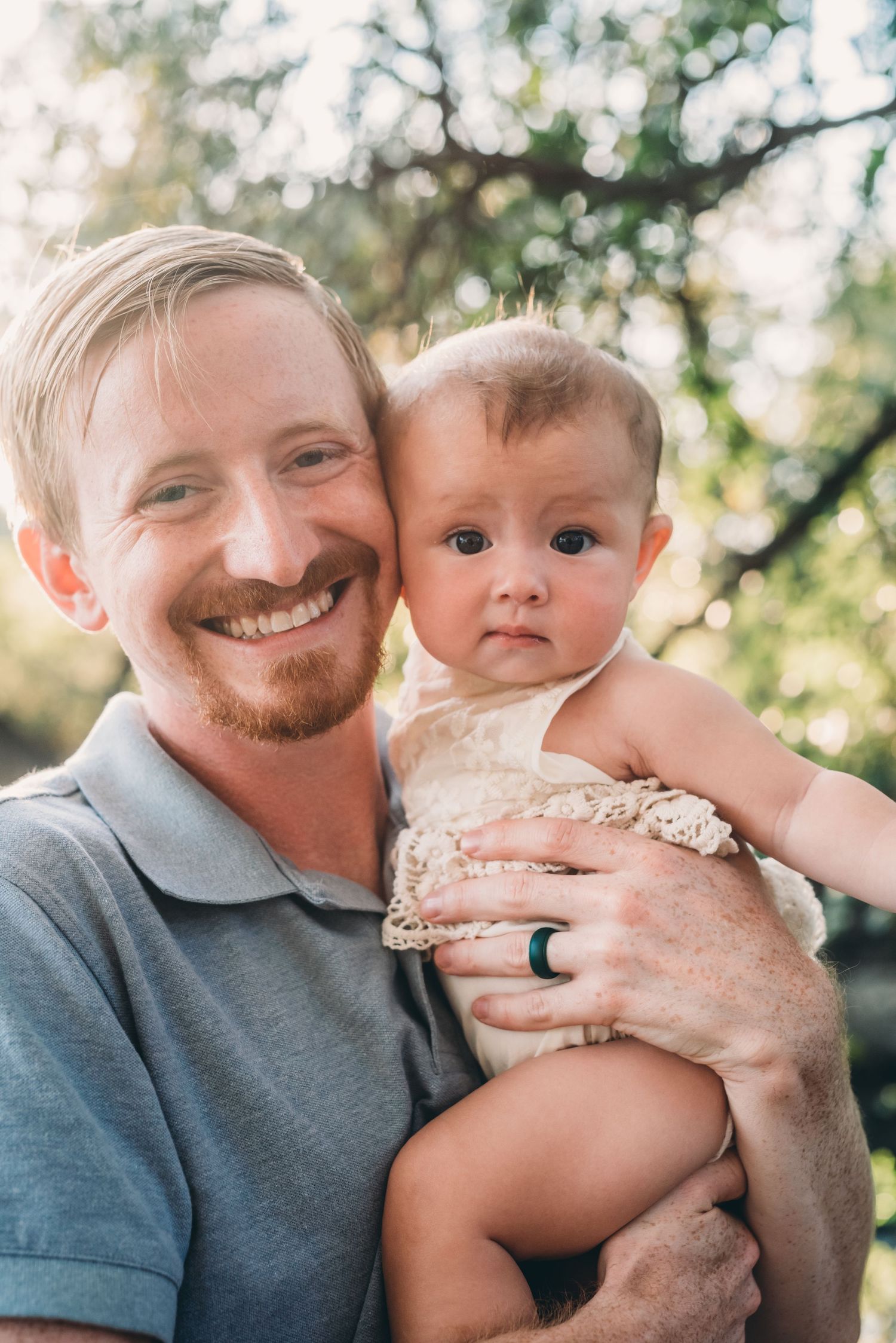Outdoor portrait of parent and baby in white lace outfit against green foliage.