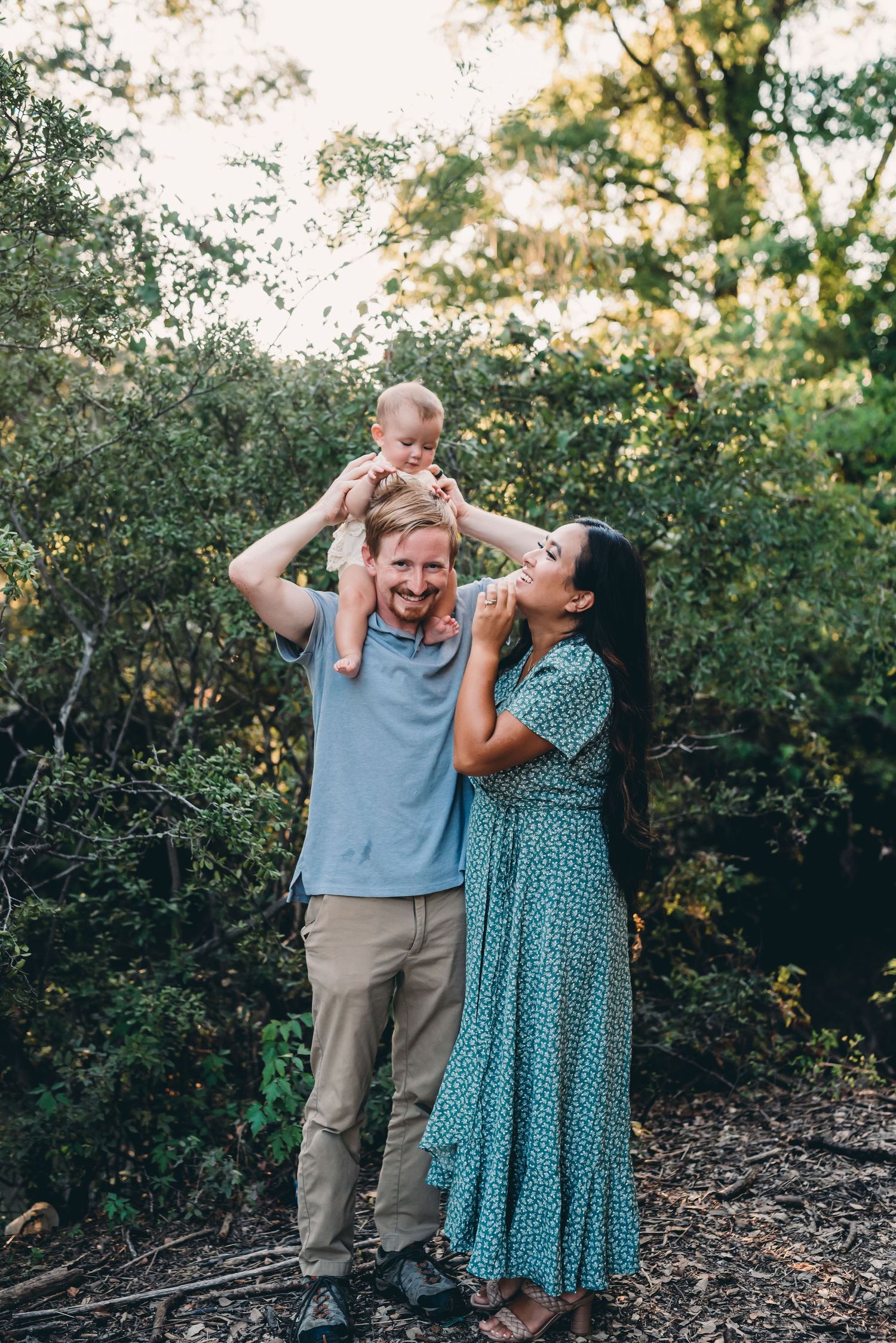 Family enjoying playful moment outdoors with baby sitting on shoulders during sunset photoshoot.