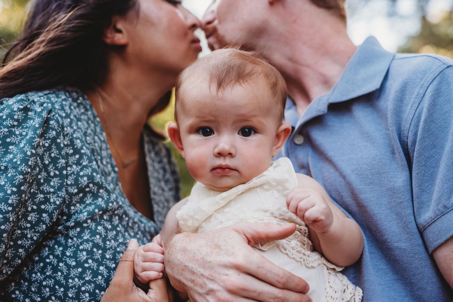 Family sharing intimate moment while holding baby in white lace outfit outdoors.