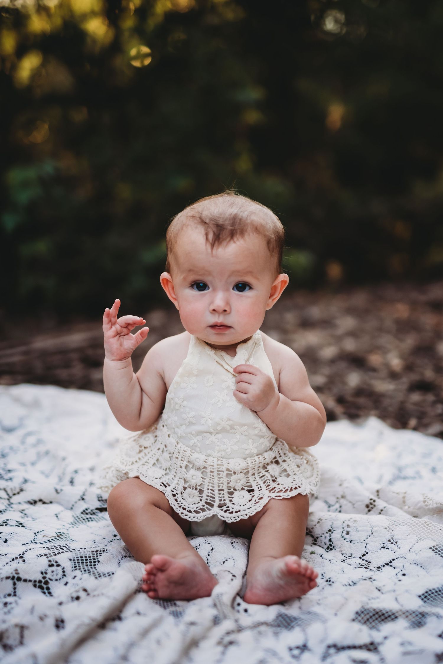 Baby in white lace dress sits on blanket outdoors.