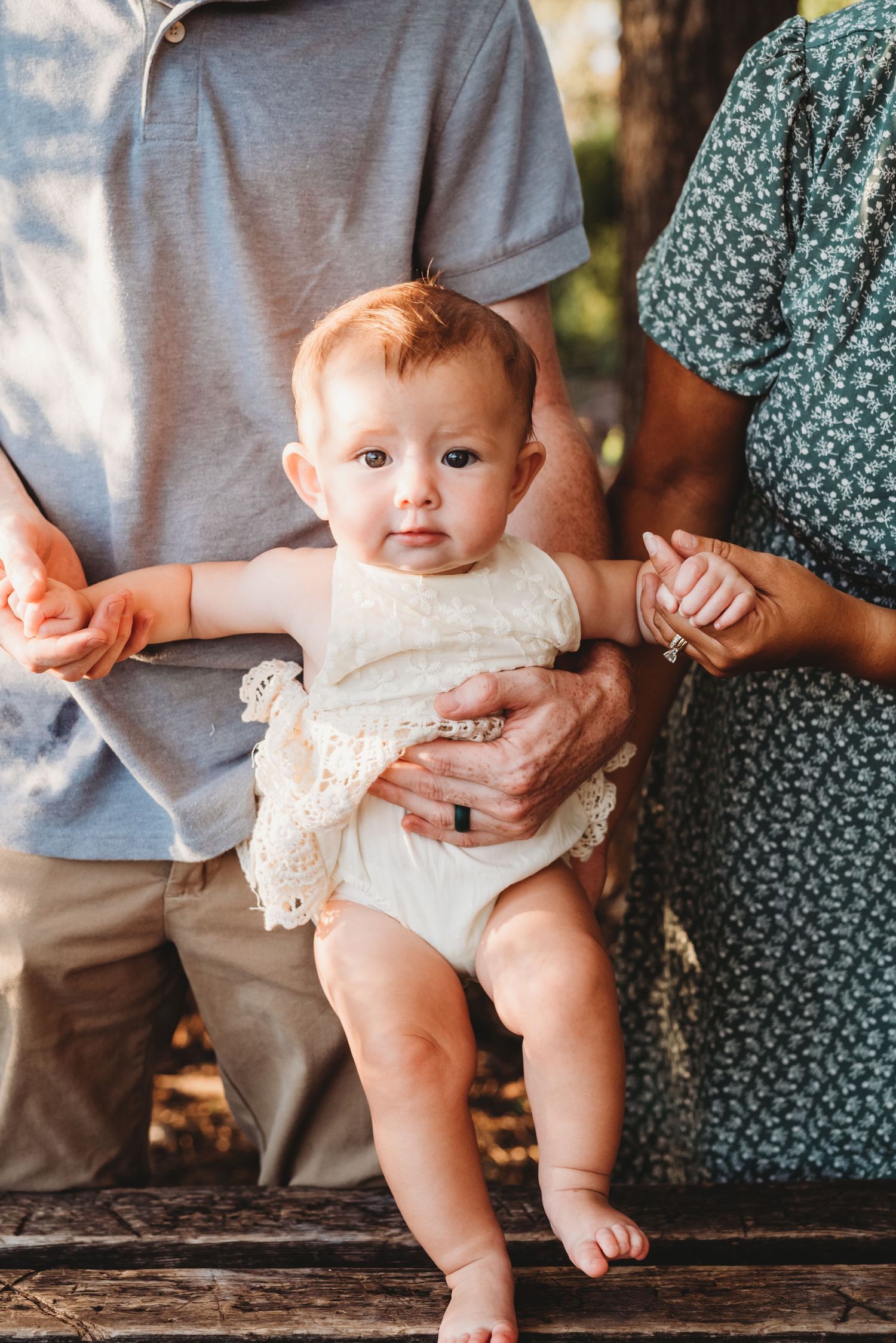 Baby in white lace outfit supported by others on wooden bench.