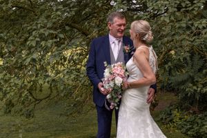 A couple poses together among green foliage with wedding flowers.