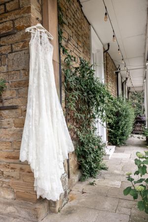 White lace wedding dress hanging on stone wall with greenery.