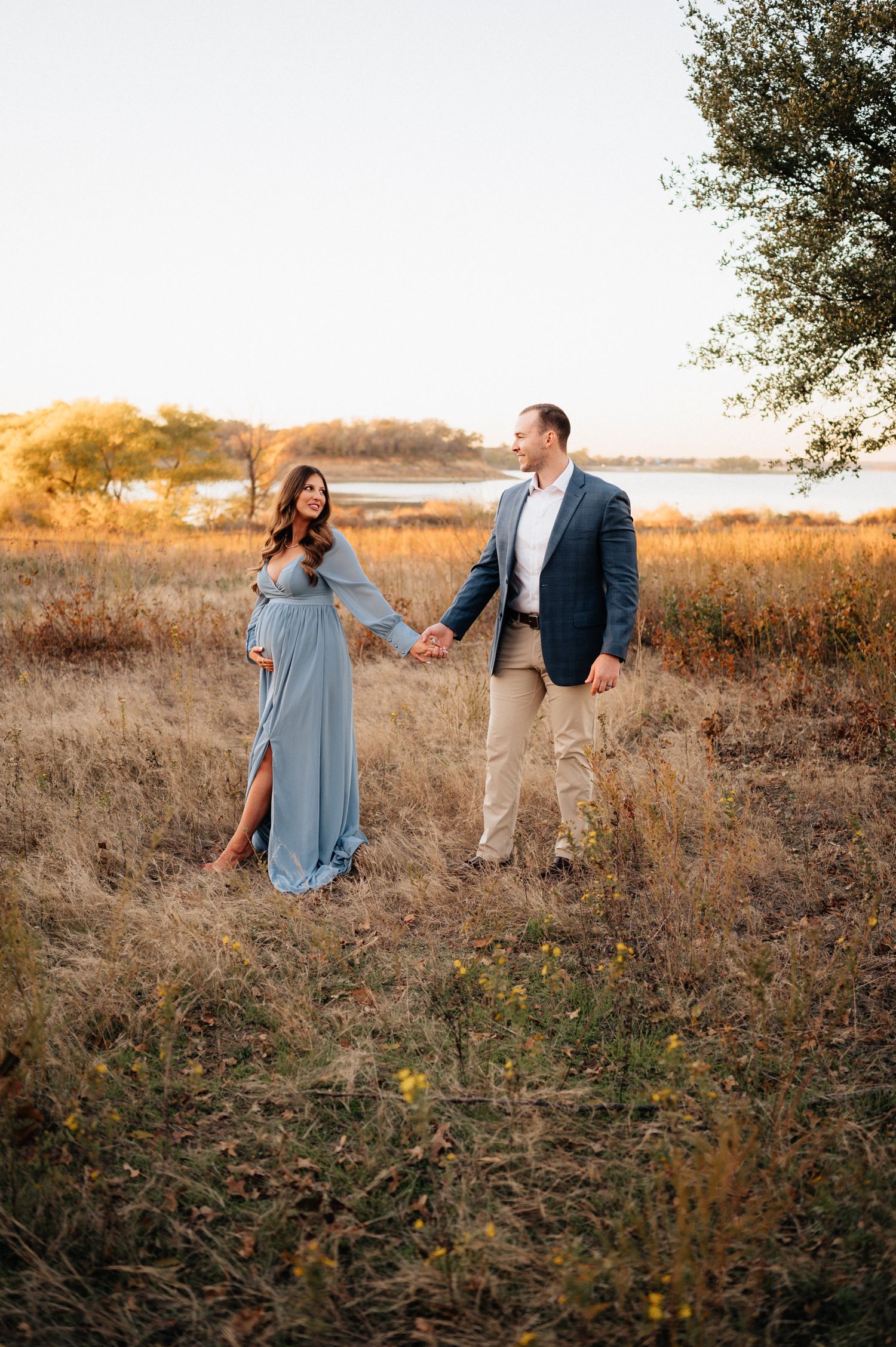 Romantic sunset engagement photo of a couple holding hands in a golden field near water.
