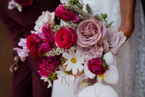 A vibrant wedding bouquet with pink roses, red ranunculus, and white daisies against a dark background.