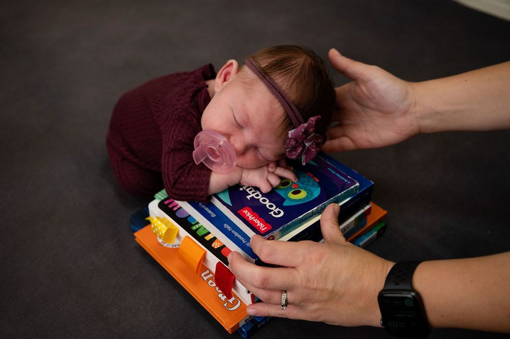Sleeping newborn rests on a collection of colorful books while hands gently support.