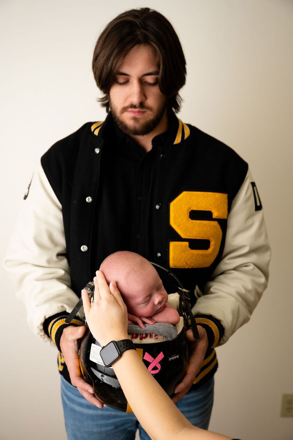 Person in black and gold varsity jacket holding sleeping newborn with hand supporting head.