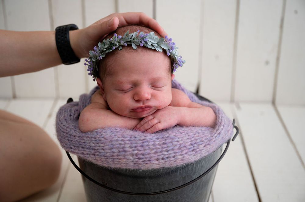 Newborn wearing floral crown sleeps peacefully in grey bucket on white wooden floor.