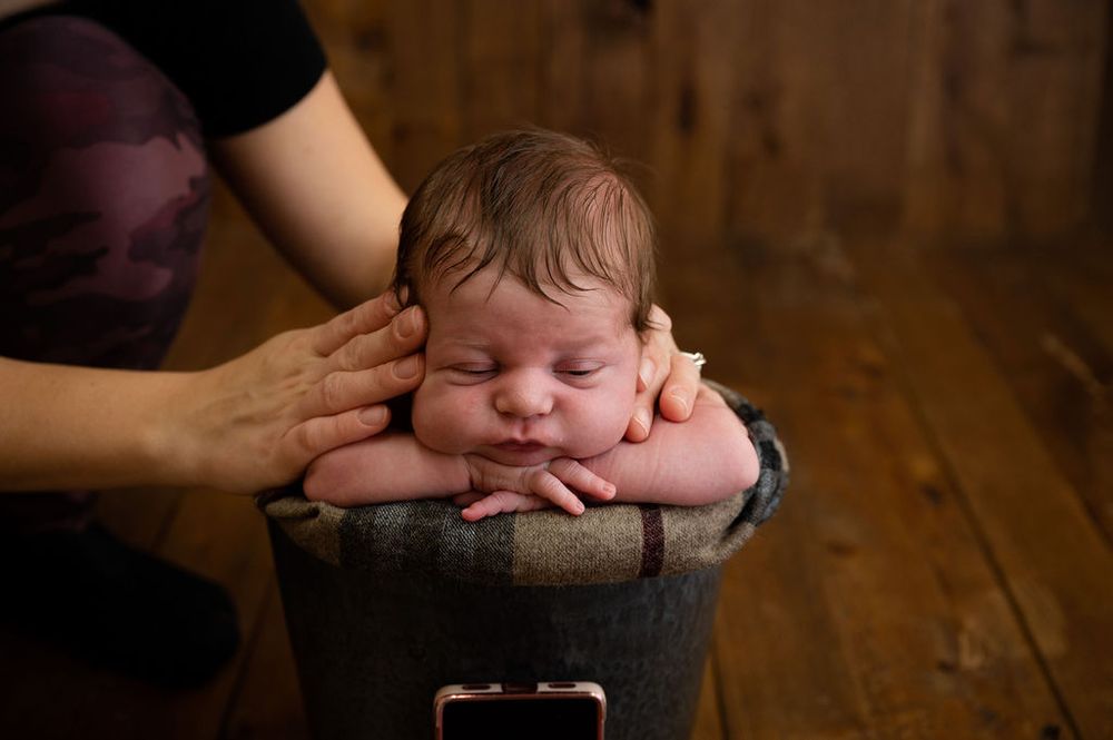 Sleeping baby rests in bucket while hands gently cradle face on wooden floor.