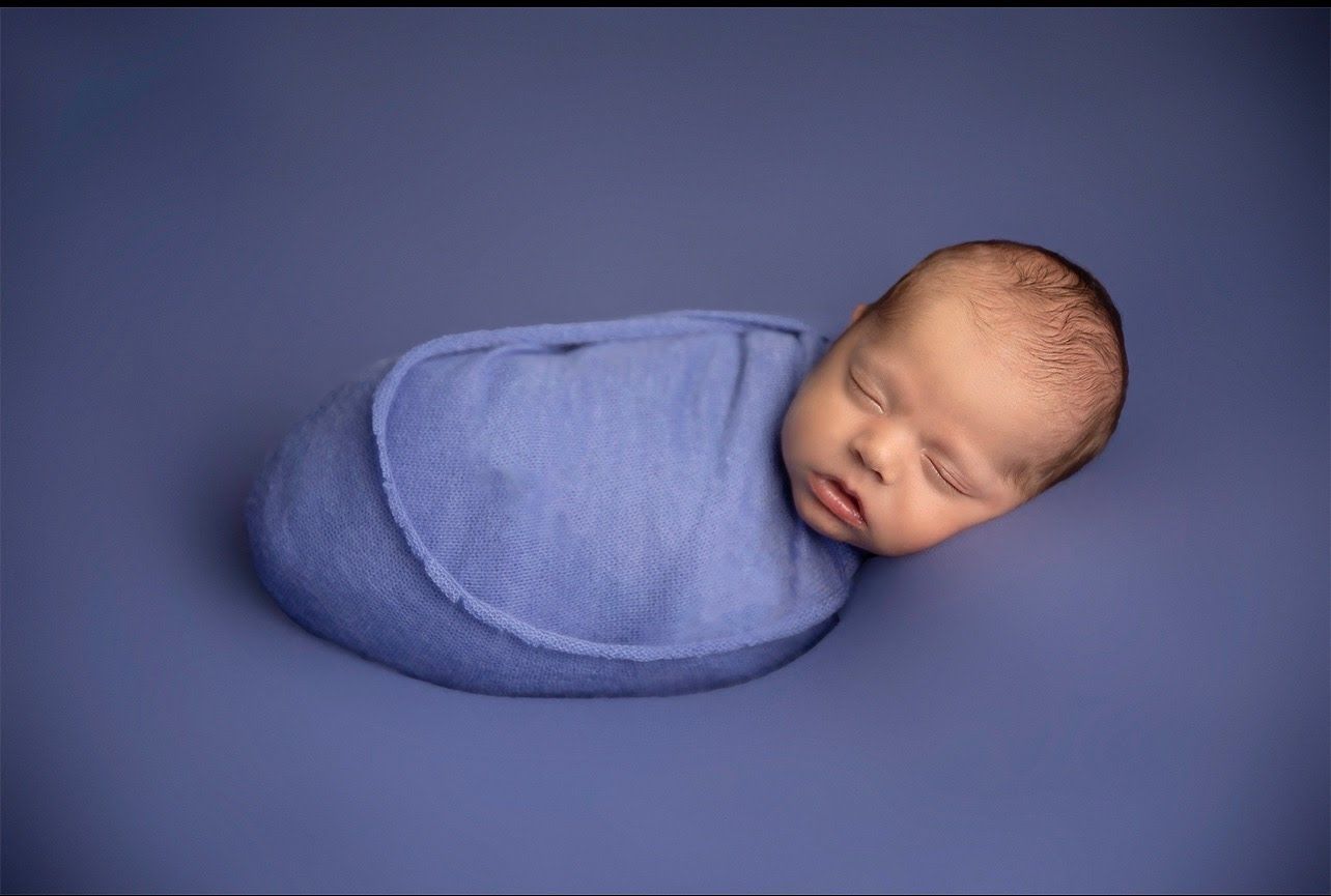 Newborn photography with baby wrapped in soft blue swaddle against navy background.