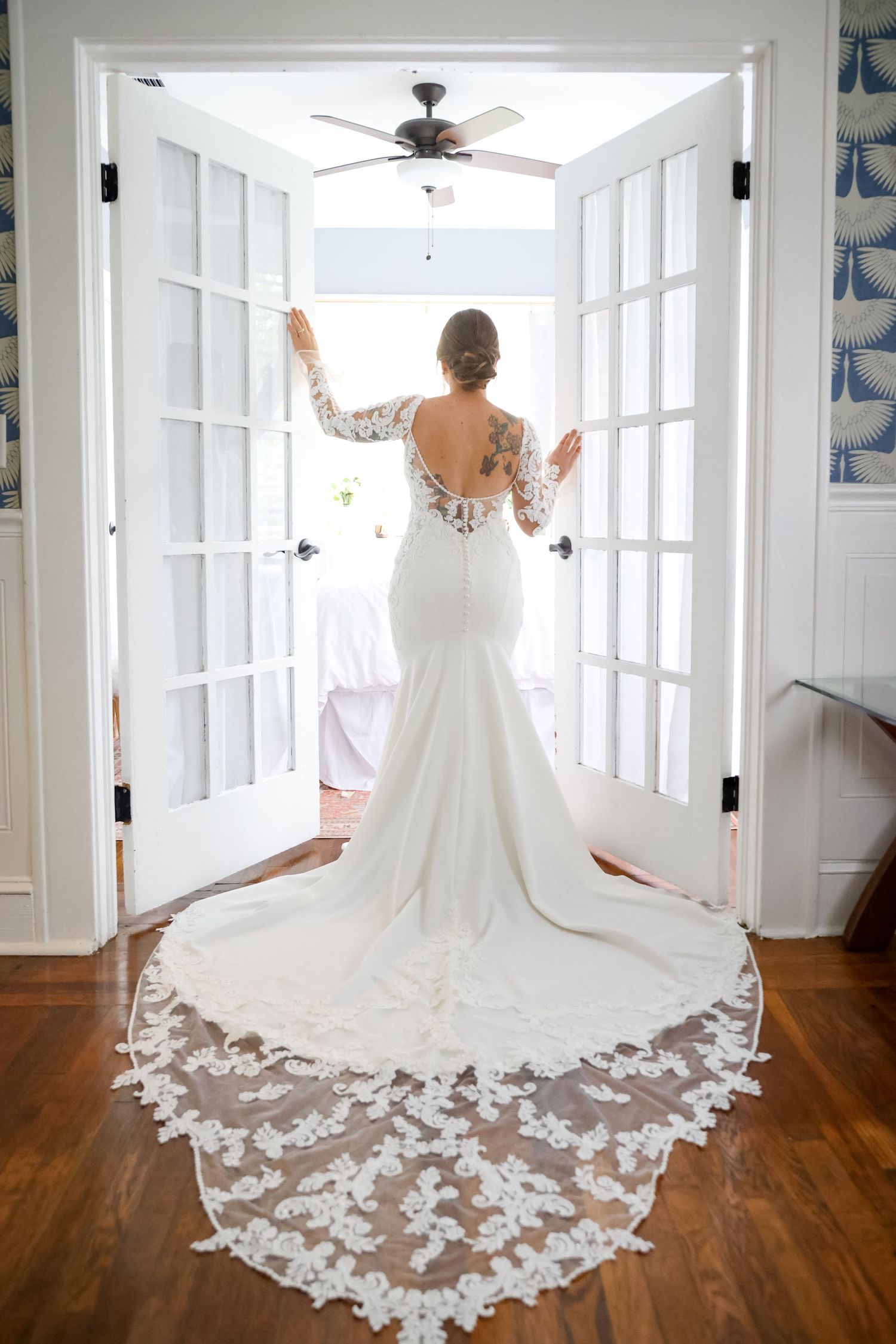 Elegant white wedding dress with backless design and dramatic lace train standing in French doorway.