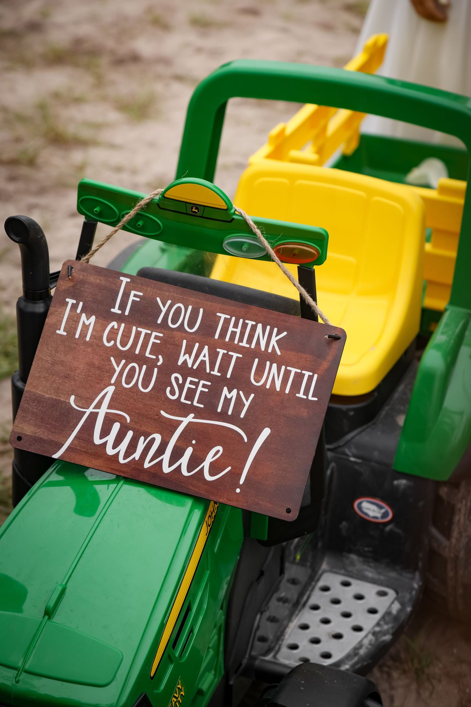 A wooden sign reading 'If you think I'm cute, wait until you see my Auntie' mounted on a green and yellow toy tractor.