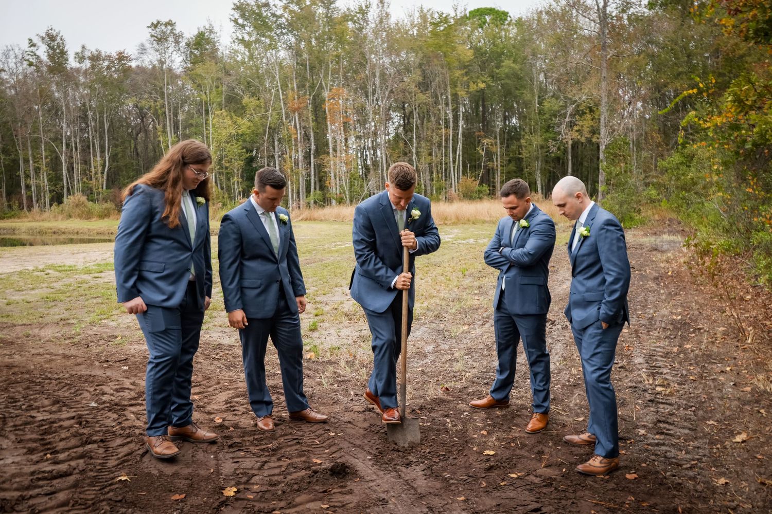 A group dressed in navy blue suits stand together on a dirt clearing near autumn trees in the background.