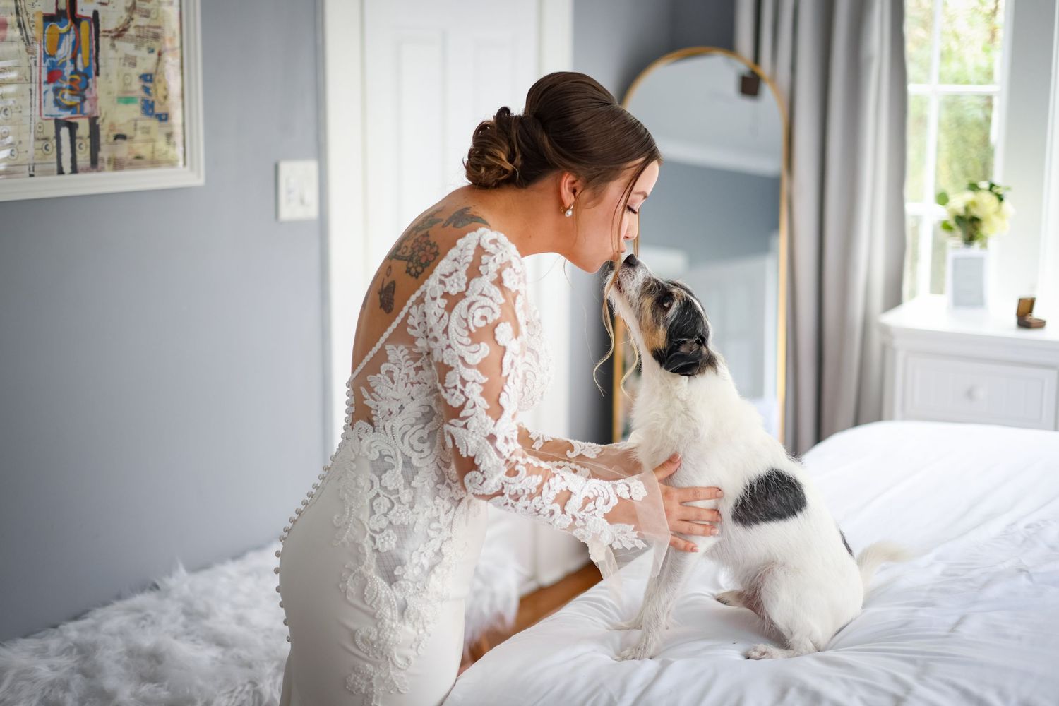 A black and white dog sits on a bed kissing a bride wearing a white lace dress in a bright bedroom setting.