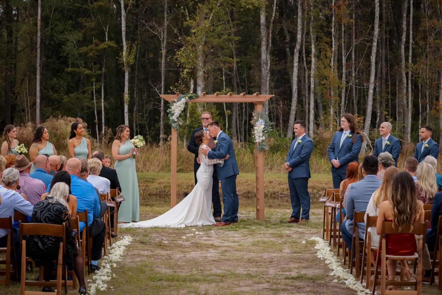 An outdoor wedding ceremony in a wooded setting with guests seated on wooden chairs and a wooden arch decorated with flowers.