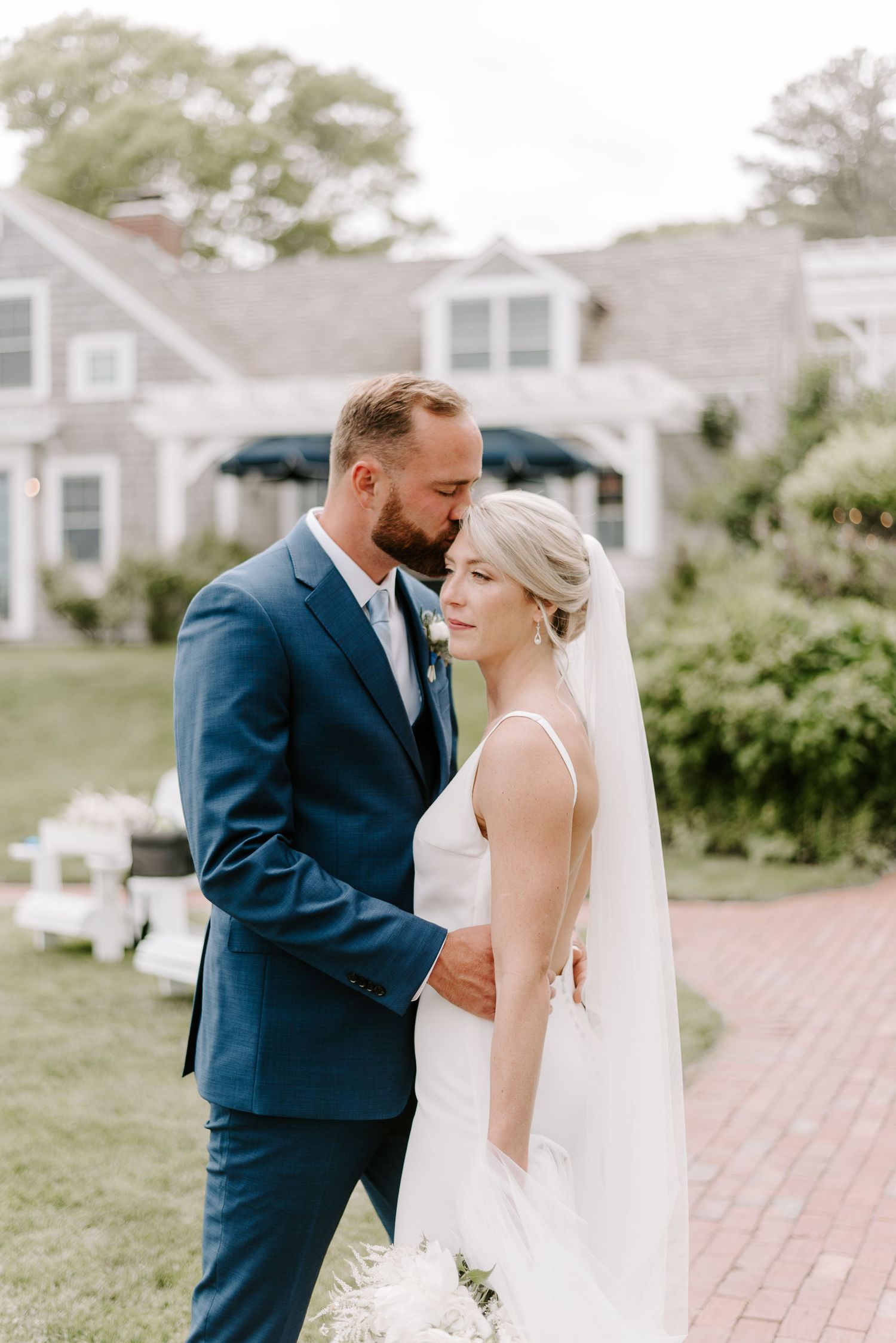 Elegant wedding couple shares an intimate moment on a brick pathway beside a classic New England home.