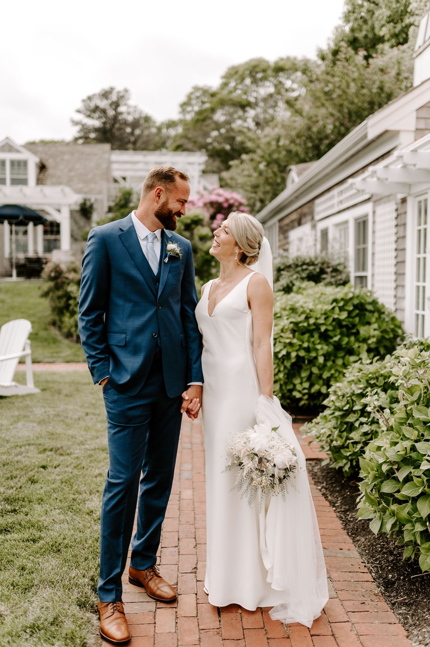 Wedding couple shares loving glance on brick walkway beside white colonial building.