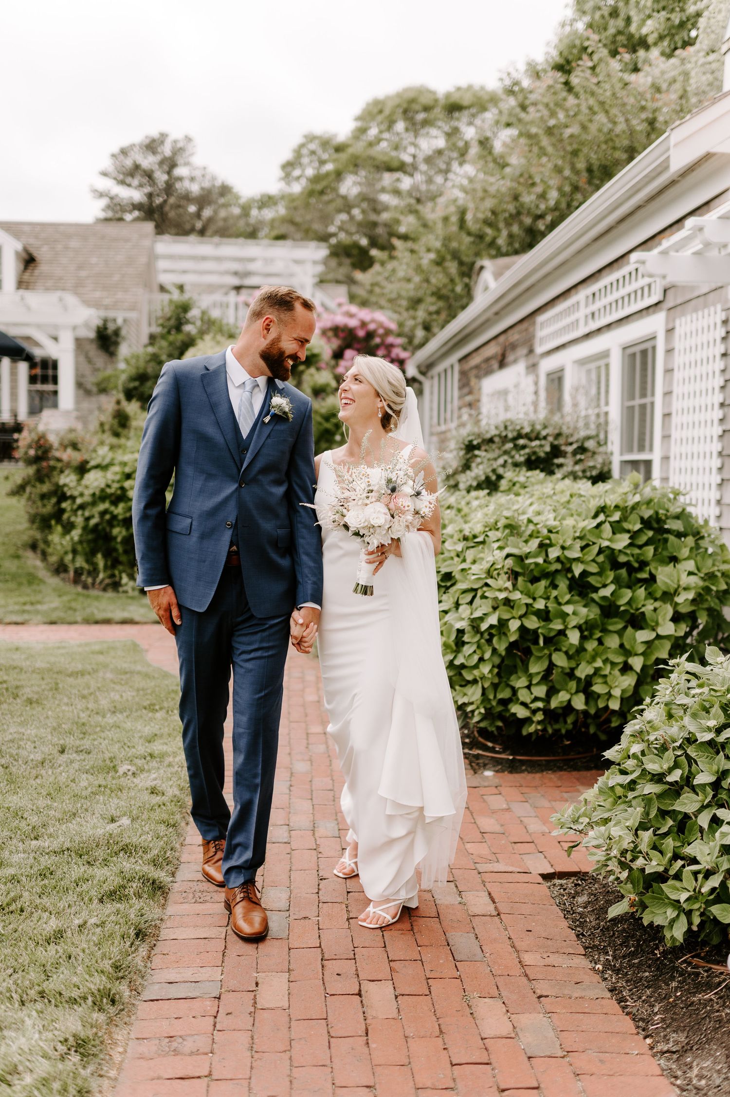 Newlyweds share a joyful moment while walking along a brick pathway surrounded by lush gardens.
