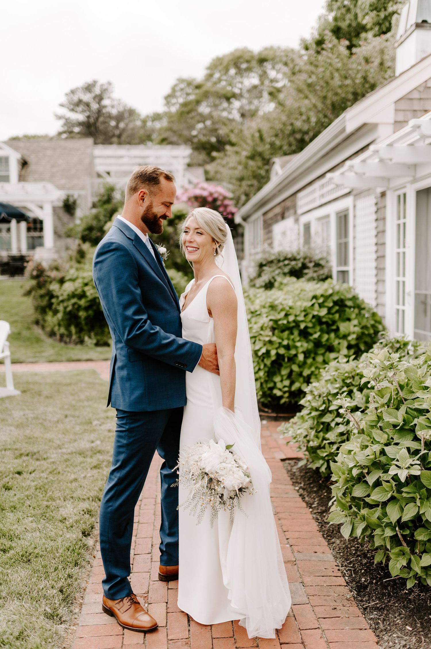 Wedding couple stands together on brick walkway beside white colonial-style building.
