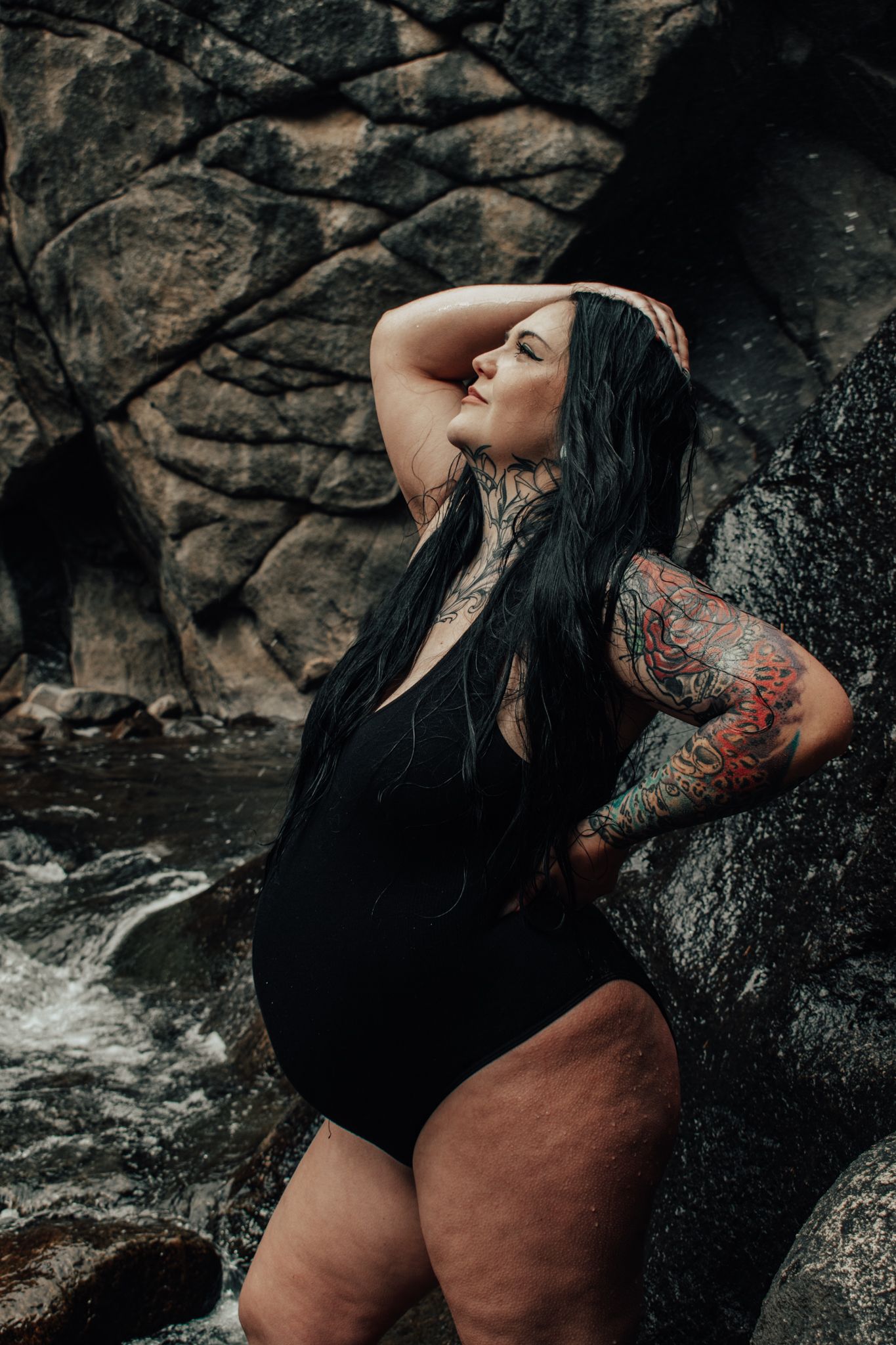 Tattooed woman in black swimsuit posing dramatically against rocky waterfall backdrop.