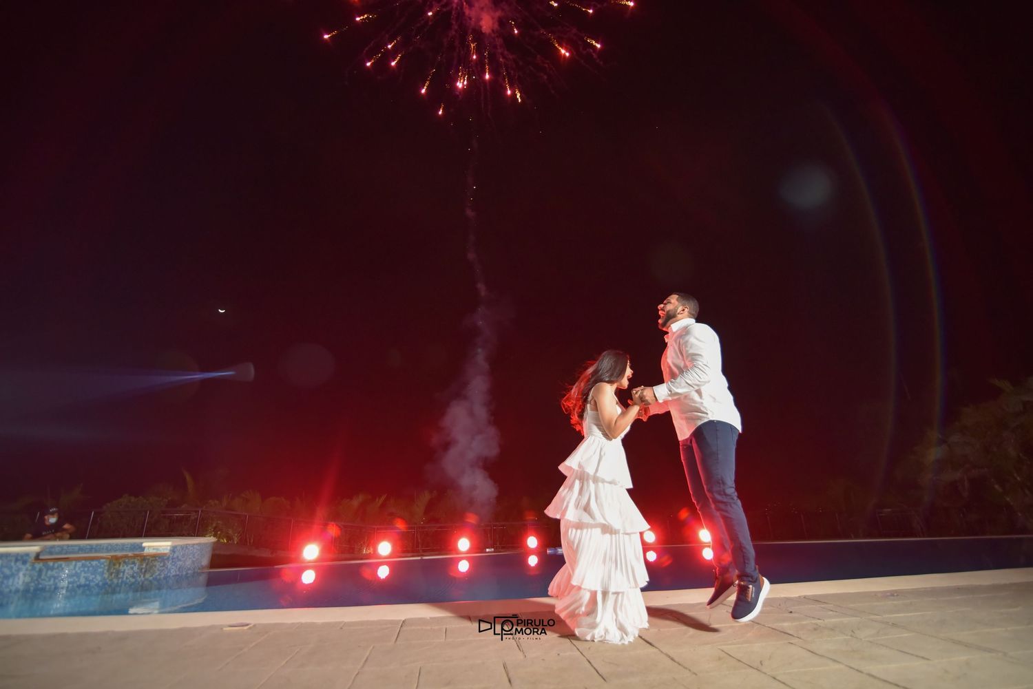 Couple dancing under colorful fireworks display at night by a pool during a romantic outdoor celebration.