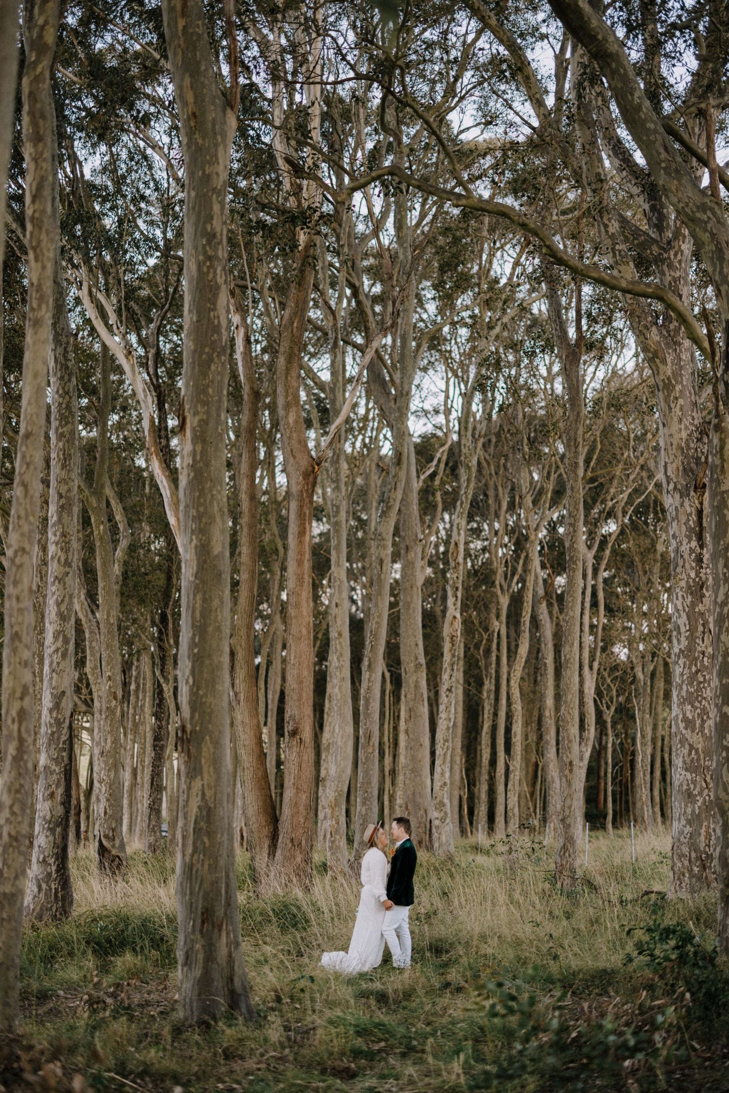 Bride and groom share a tender moment surrounded by tall trees in a forest.