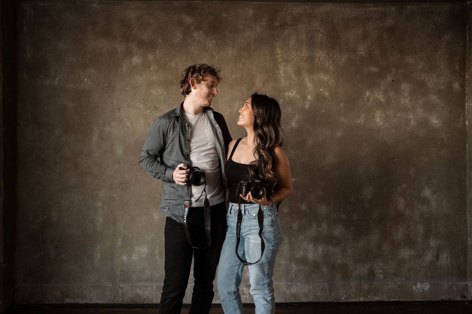 Two photographers with cameras standing against an industrial concrete wall.