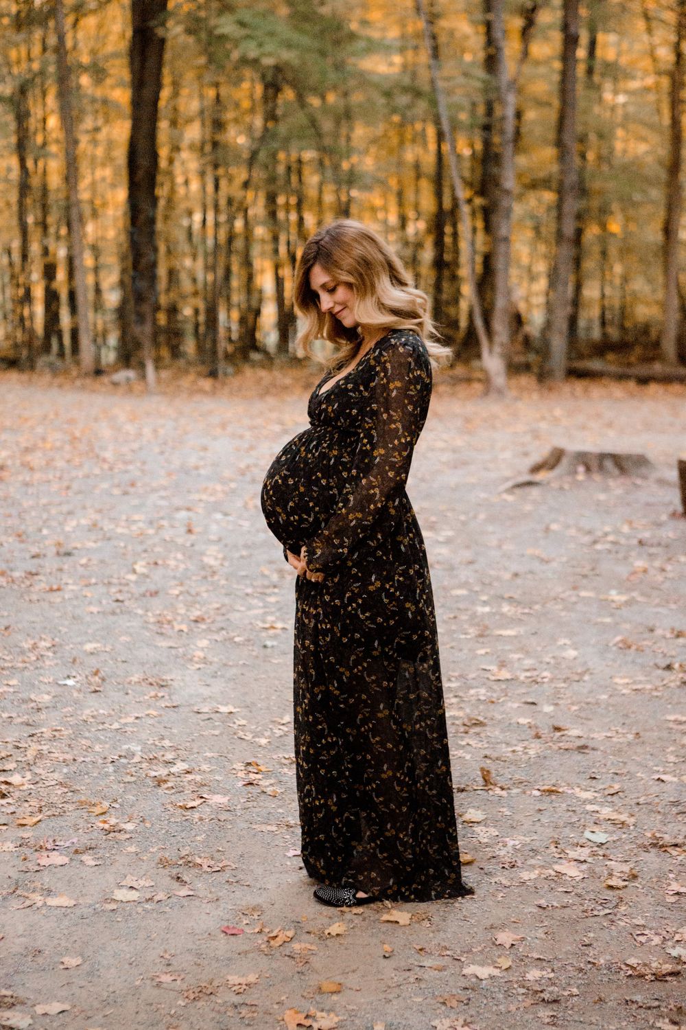 Full-length maternity portrait in black dress against autumn forest backdrop.