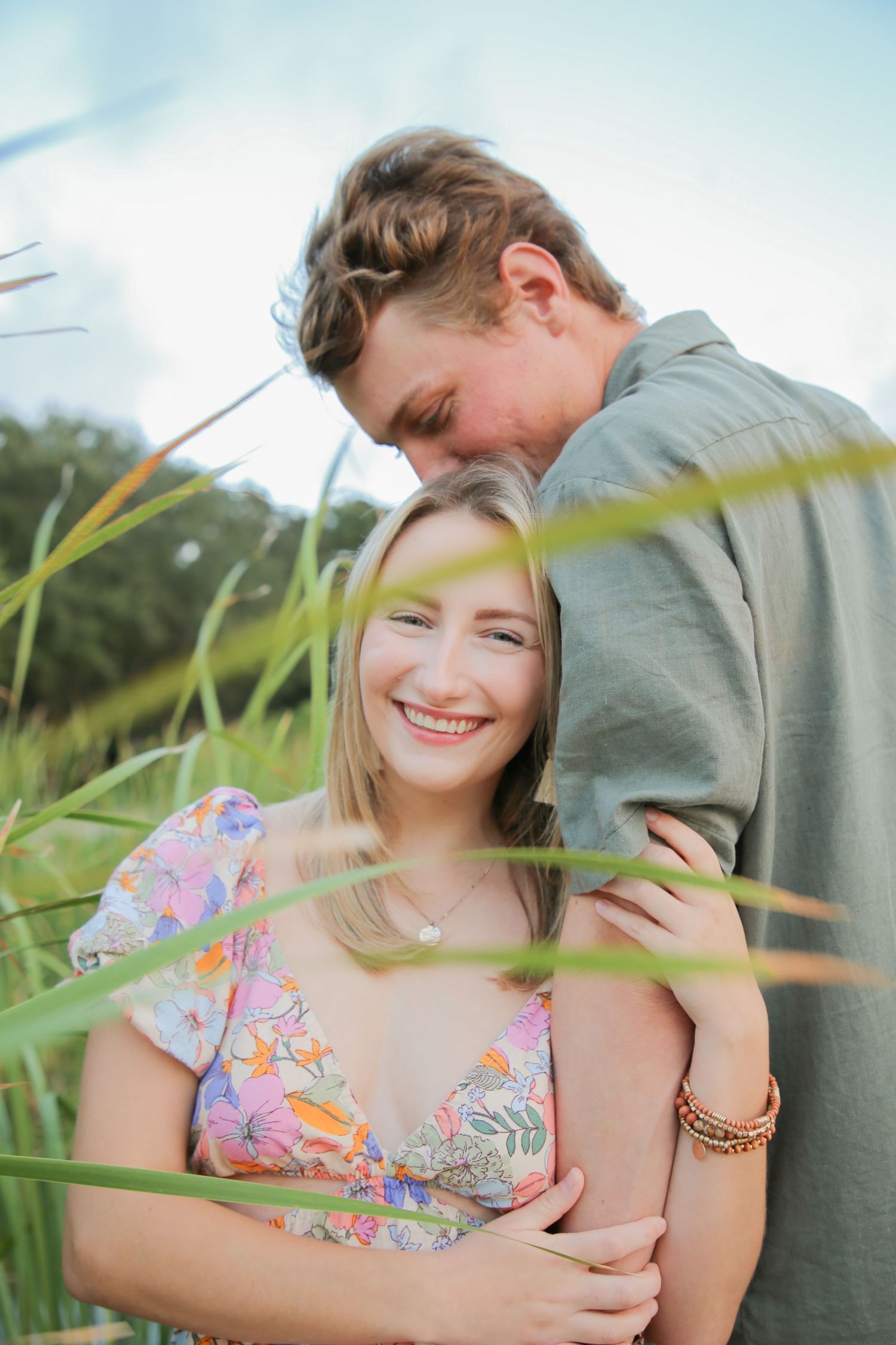 Smiling couple embracing in tall green grass, woman in floral dress laughing joyfully outdoors.