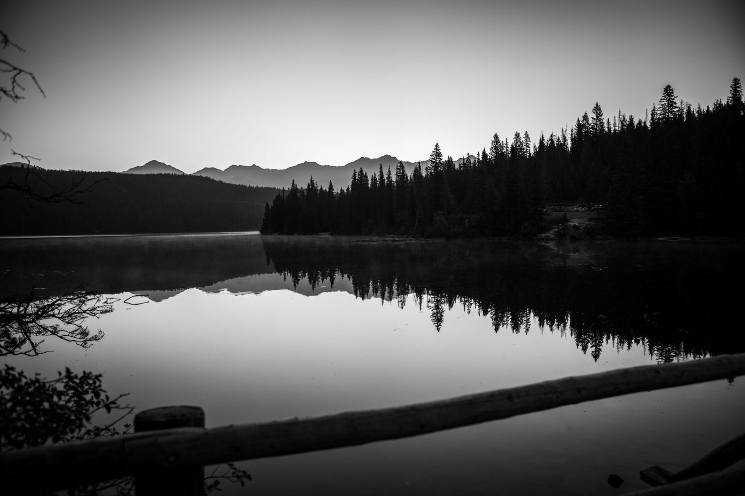 Black and white landscape of a calm mountain lake reflecting pine trees and distant peaks.