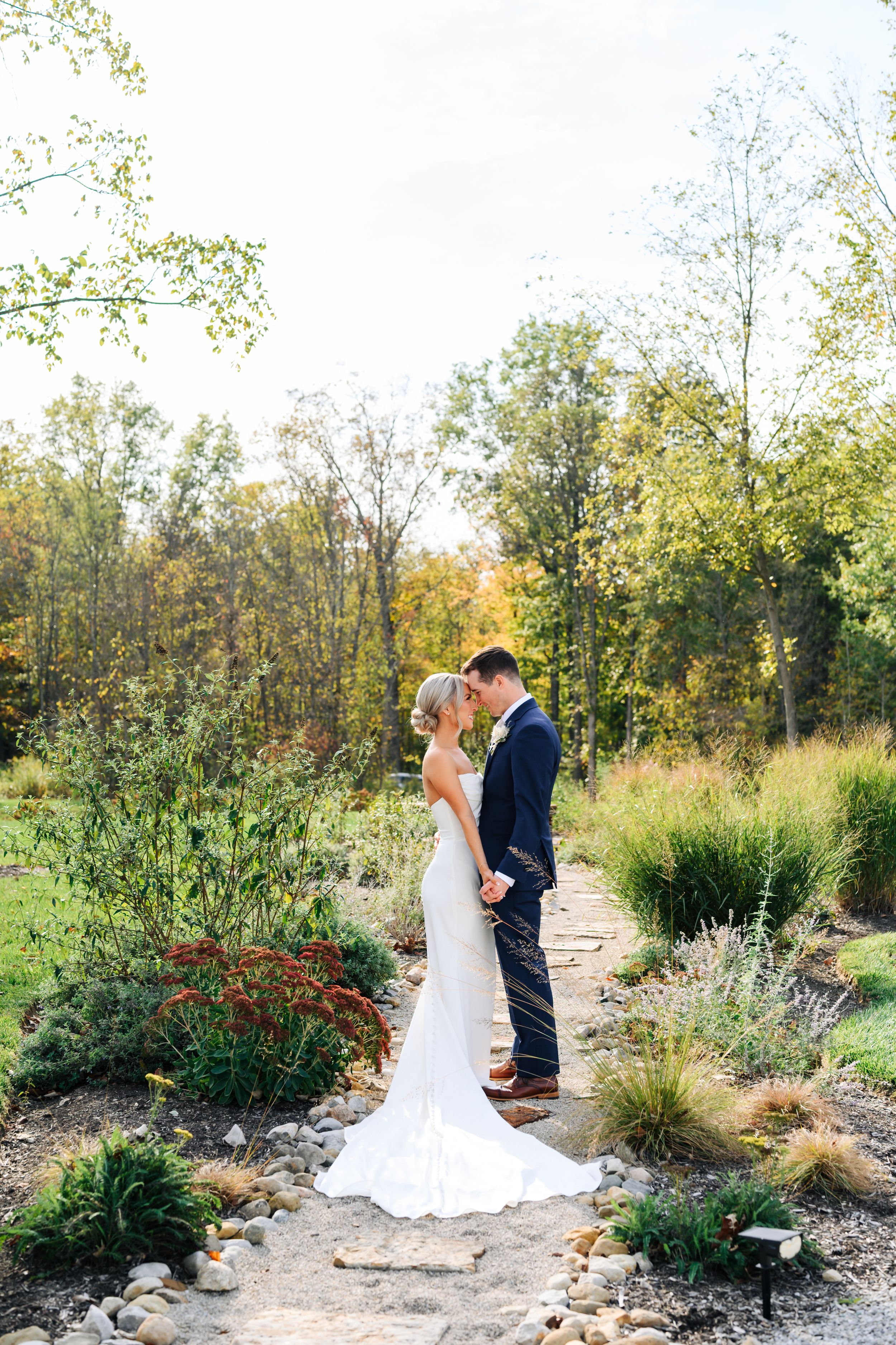 A couple shares an intimate moment on a garden path surrounded by autumn foliage and ornamental grasses.
