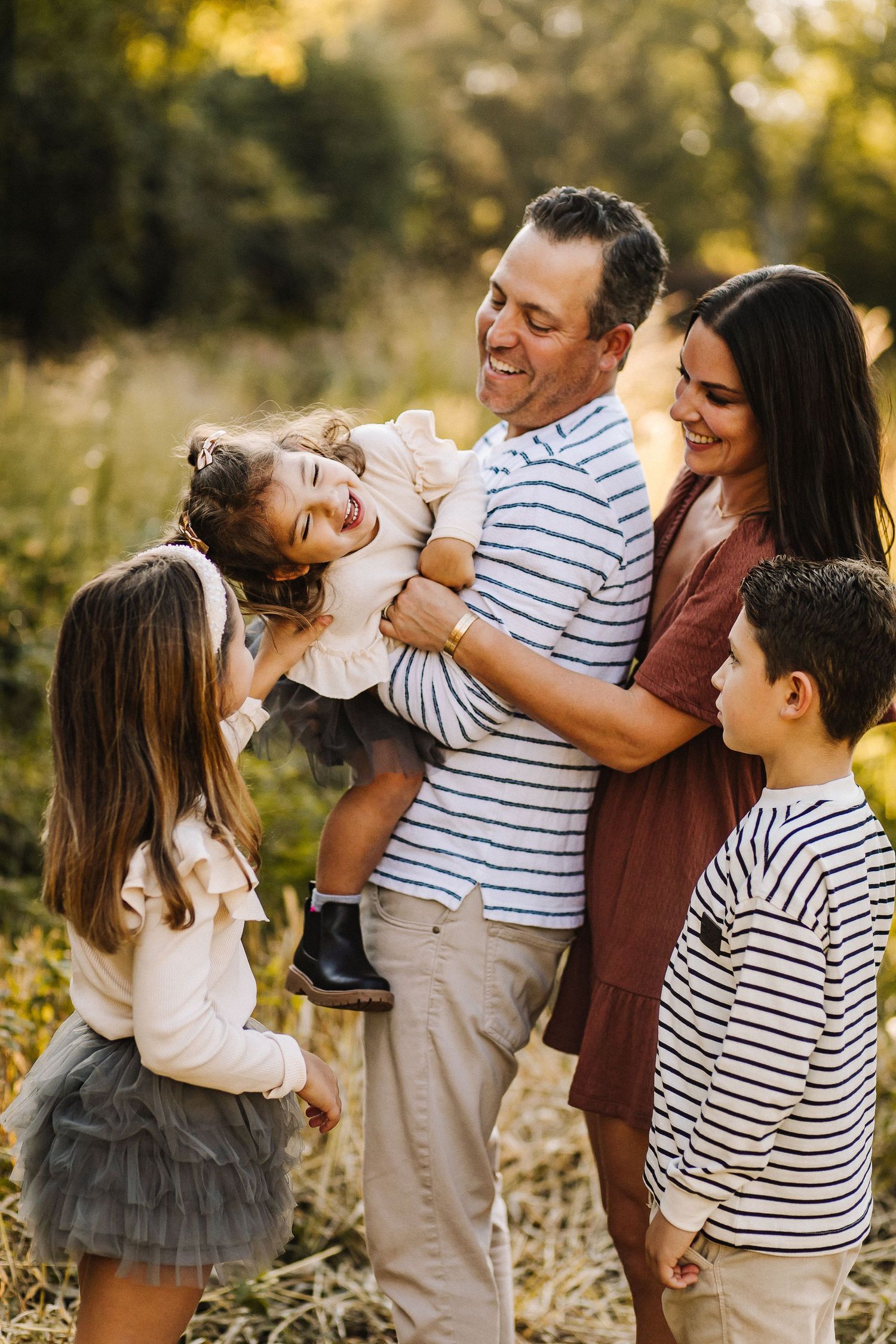 Family of five sharing laughs and cuddles together in a sunlit field at golden hour.