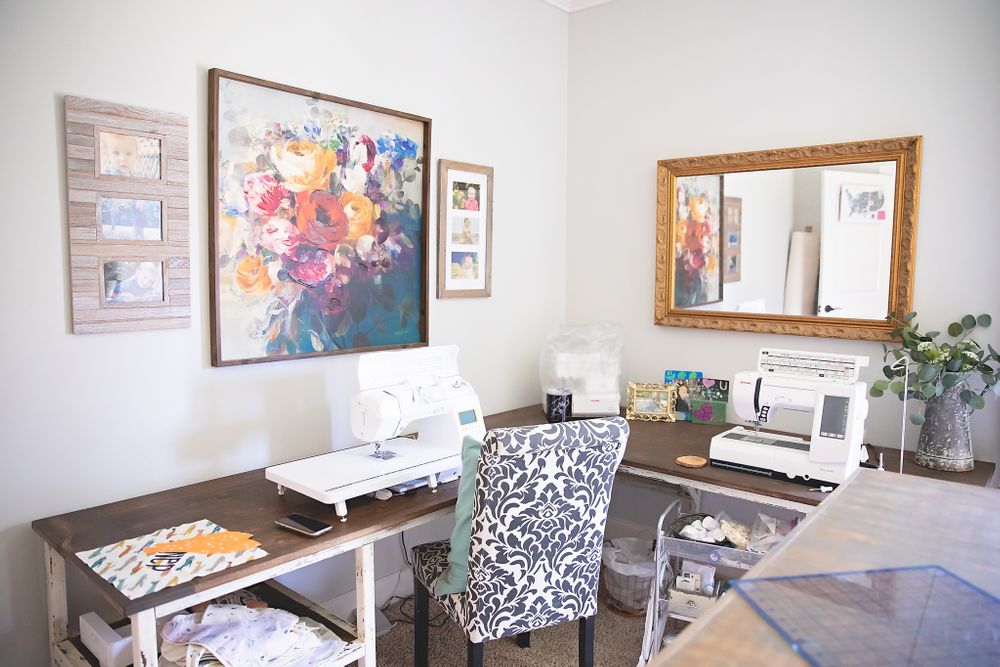 A home office workspace with a patterned chair, framed artwork, and sewing machines on a desk.