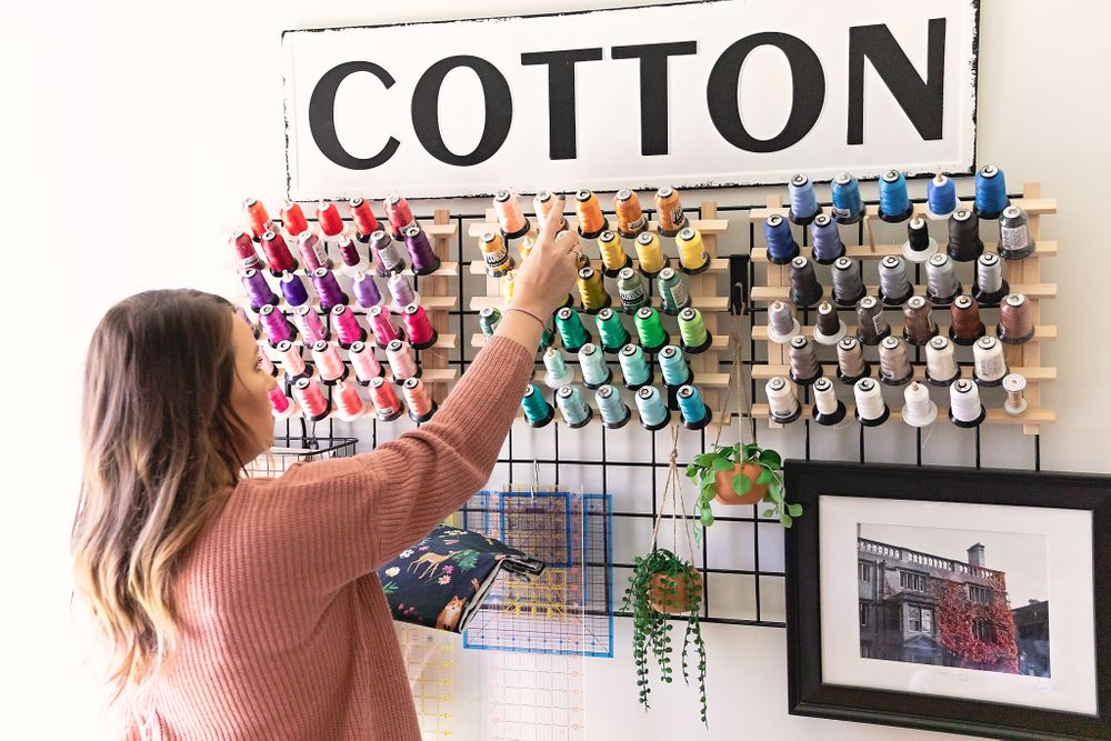 Hand reaching for spools of colorful thread on a wall-mounted organizer with 'COTTON' sign.