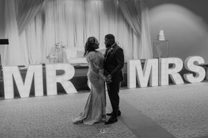A black and white photo of newlyweds posing in front of large MR & MRS letters at their reception.