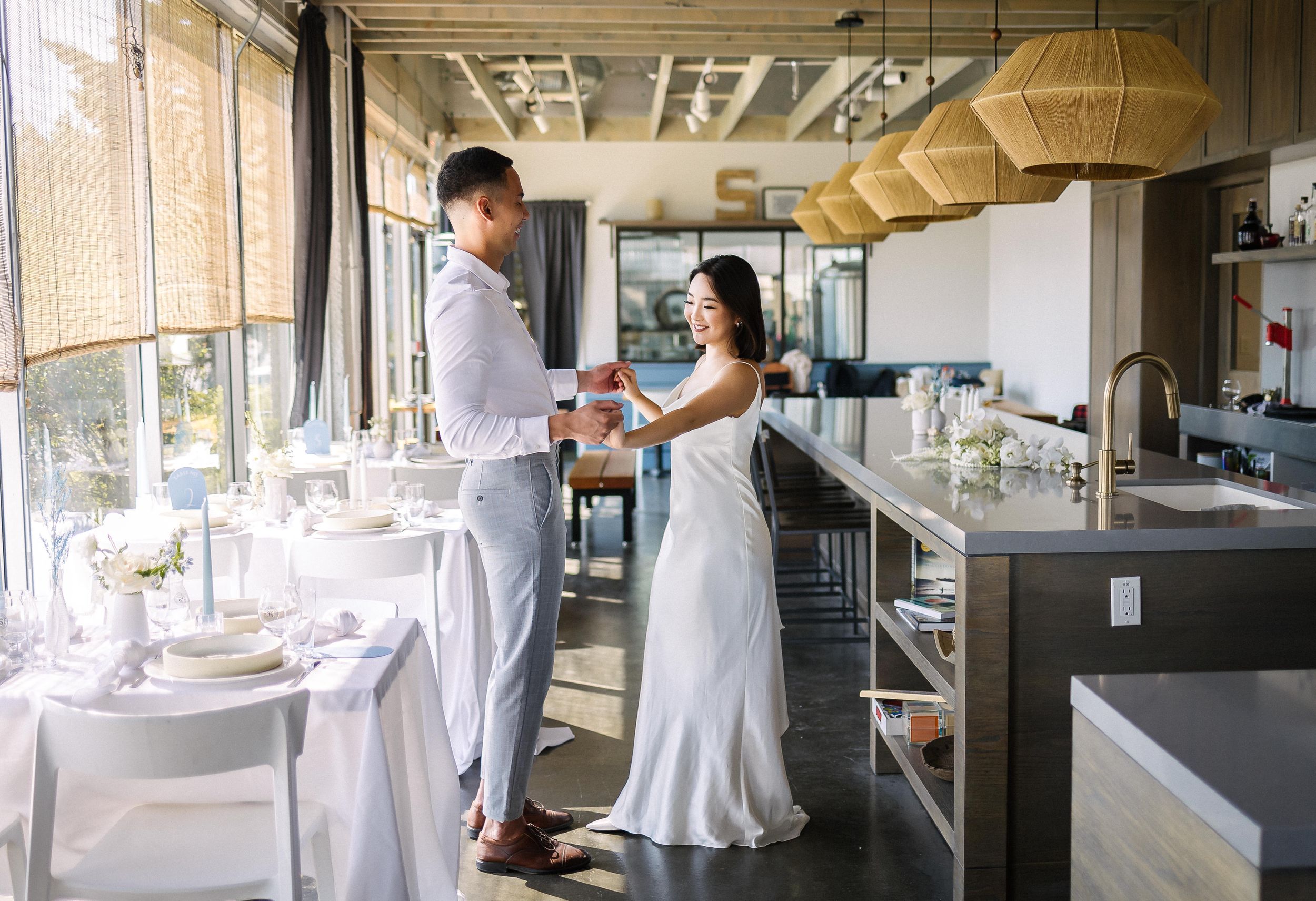 A couple in white attire share a tender moment in an elegantly set dining room.