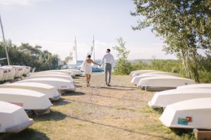 Two figures walk along a path lined with white overturned boats on a sunny summer day.
