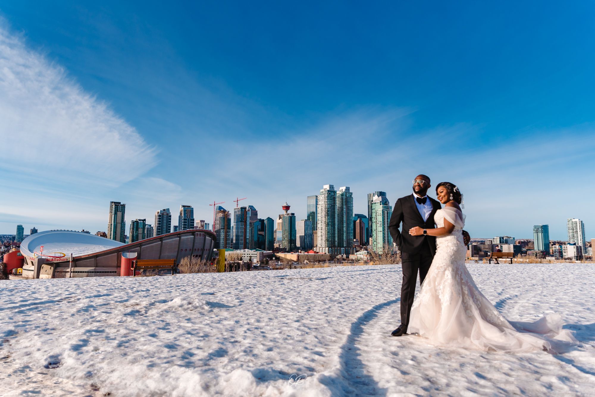 Winter wedding couple poses on snowy ground with city skyline backdrop.