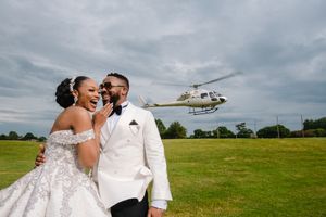 A joyful wedding couple shares a laugh on a grassy field with a helicopter in the background.