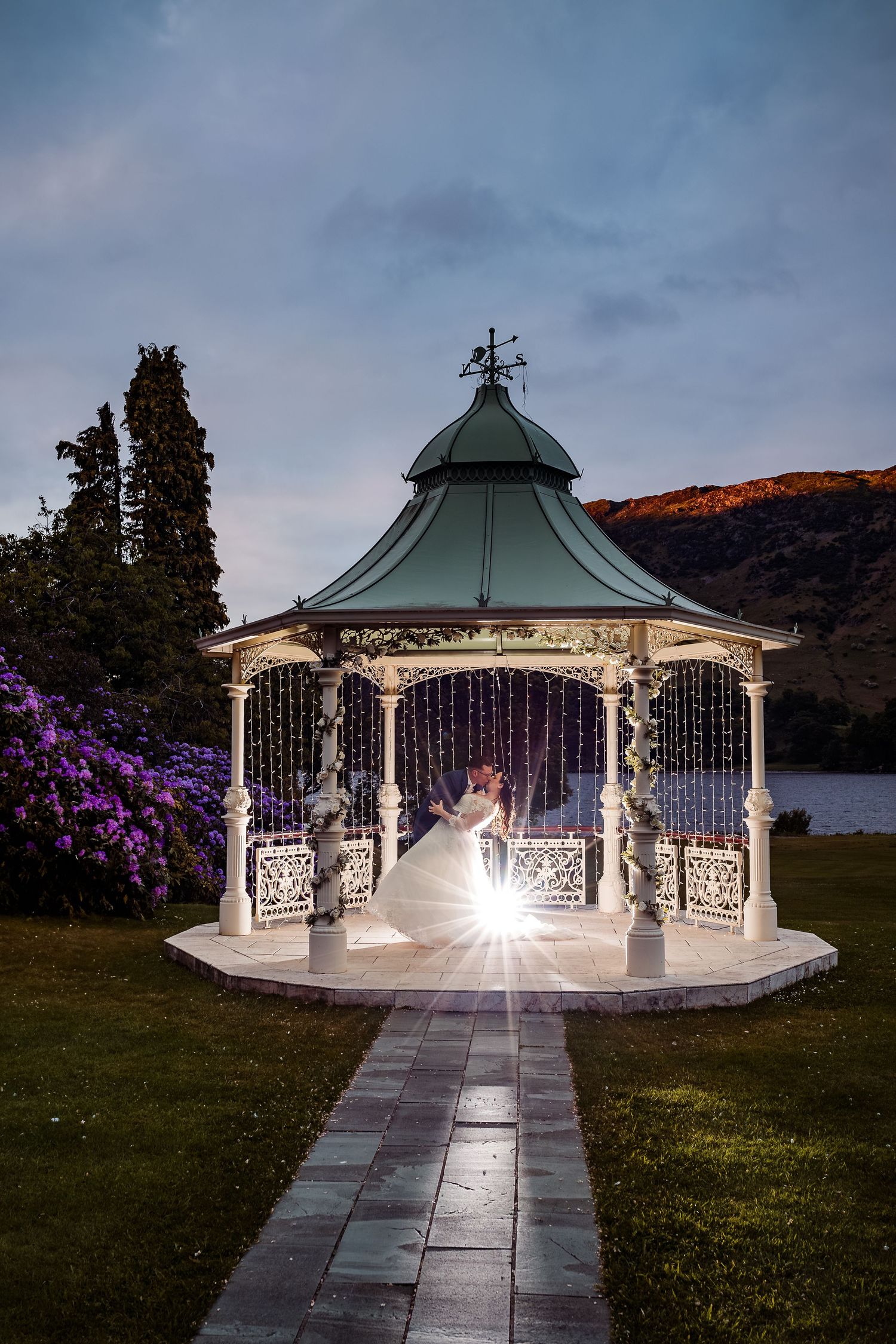 An illuminated gazebo creates a magical wedding scene at dusk with purple flowers in the foreground.