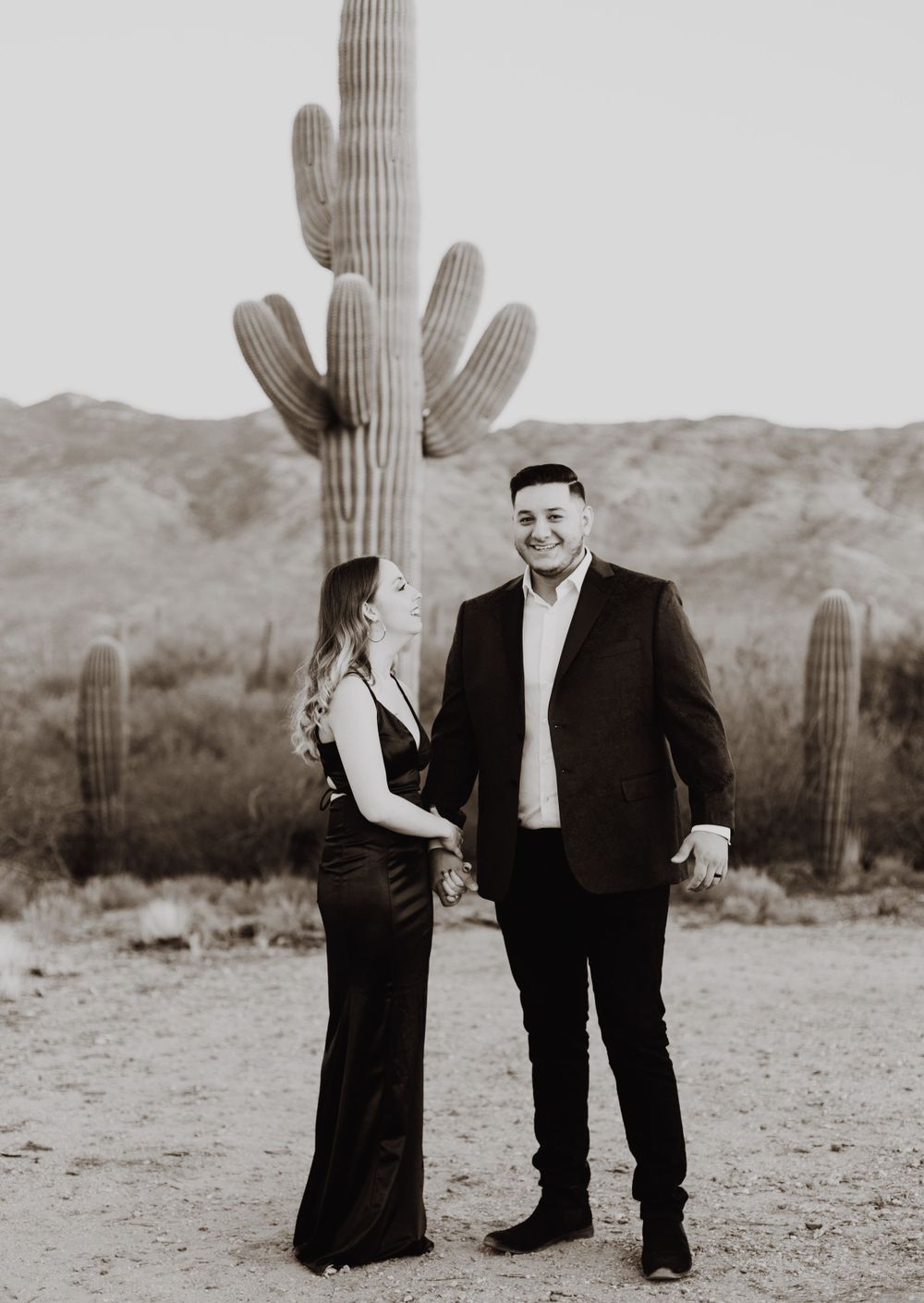 A couple poses in formal attire in front of a tall saguaro cactus in the desert.