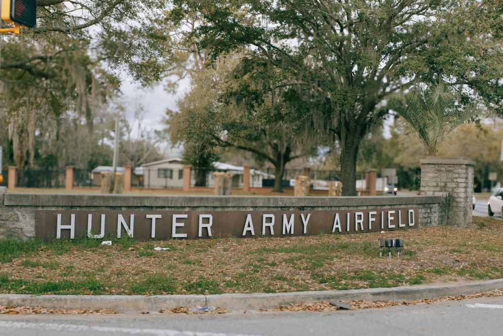 An entrance sign for Hunter Army Airfield with large white letters against a mossy southern landscape with oak trees.