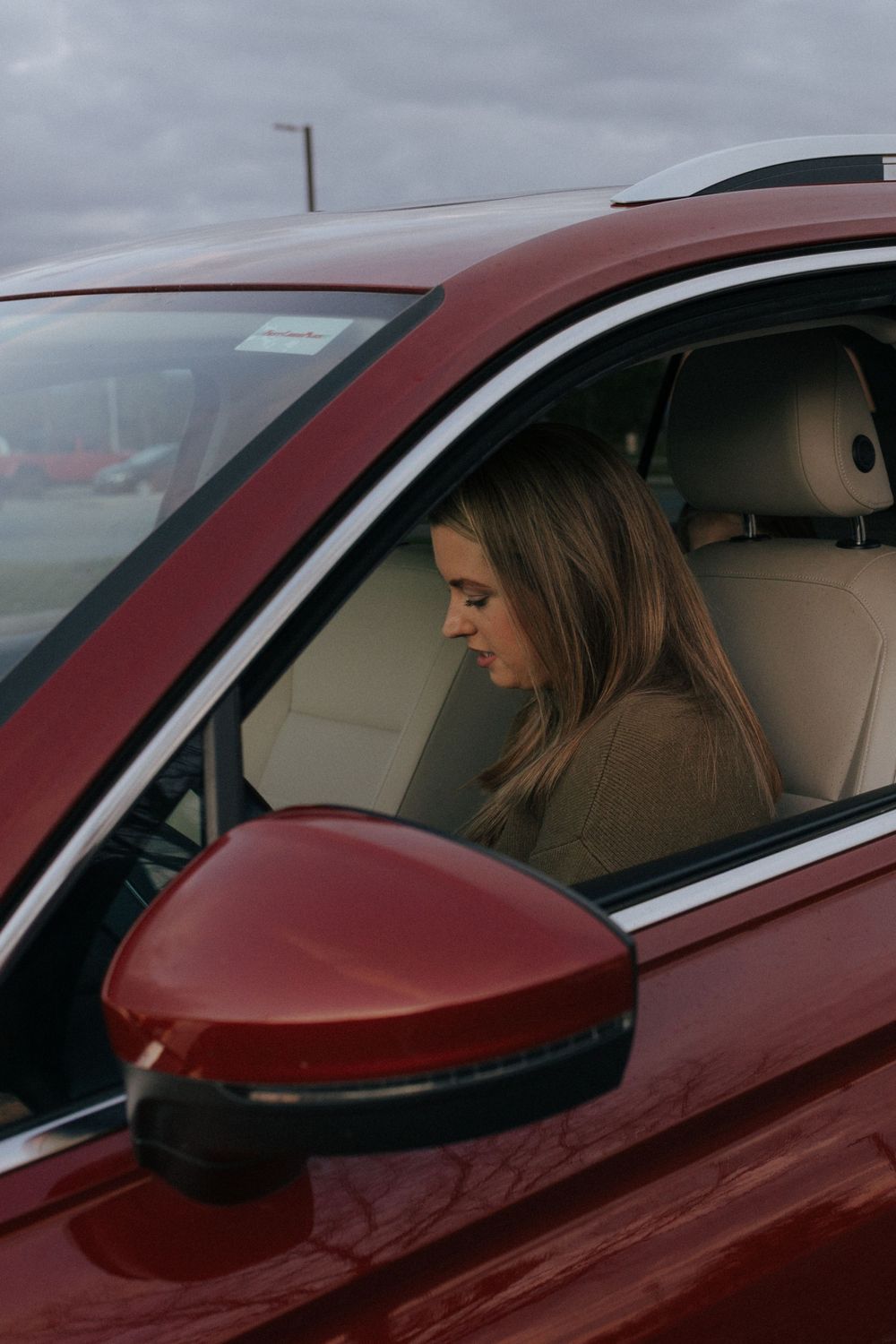 A person sits in the driver's seat of a red car with cream leather interior on a cloudy day.
