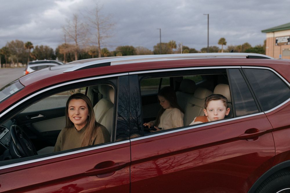 People sitting inside a red SUV on a cloudy day with buildings in the background.