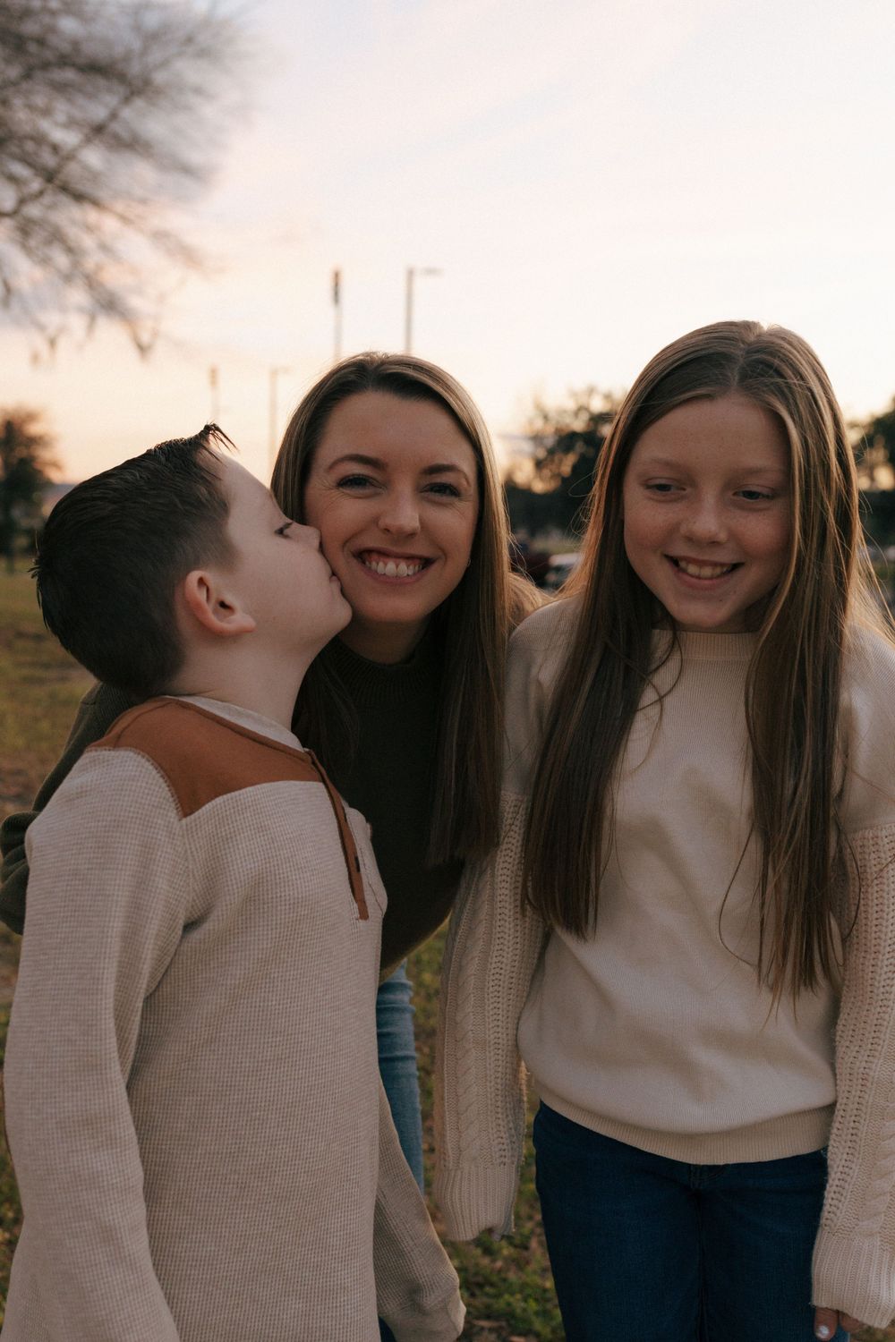 A candid outdoor family portrait at sunset showing joyful interaction between three people wearing sweaters.