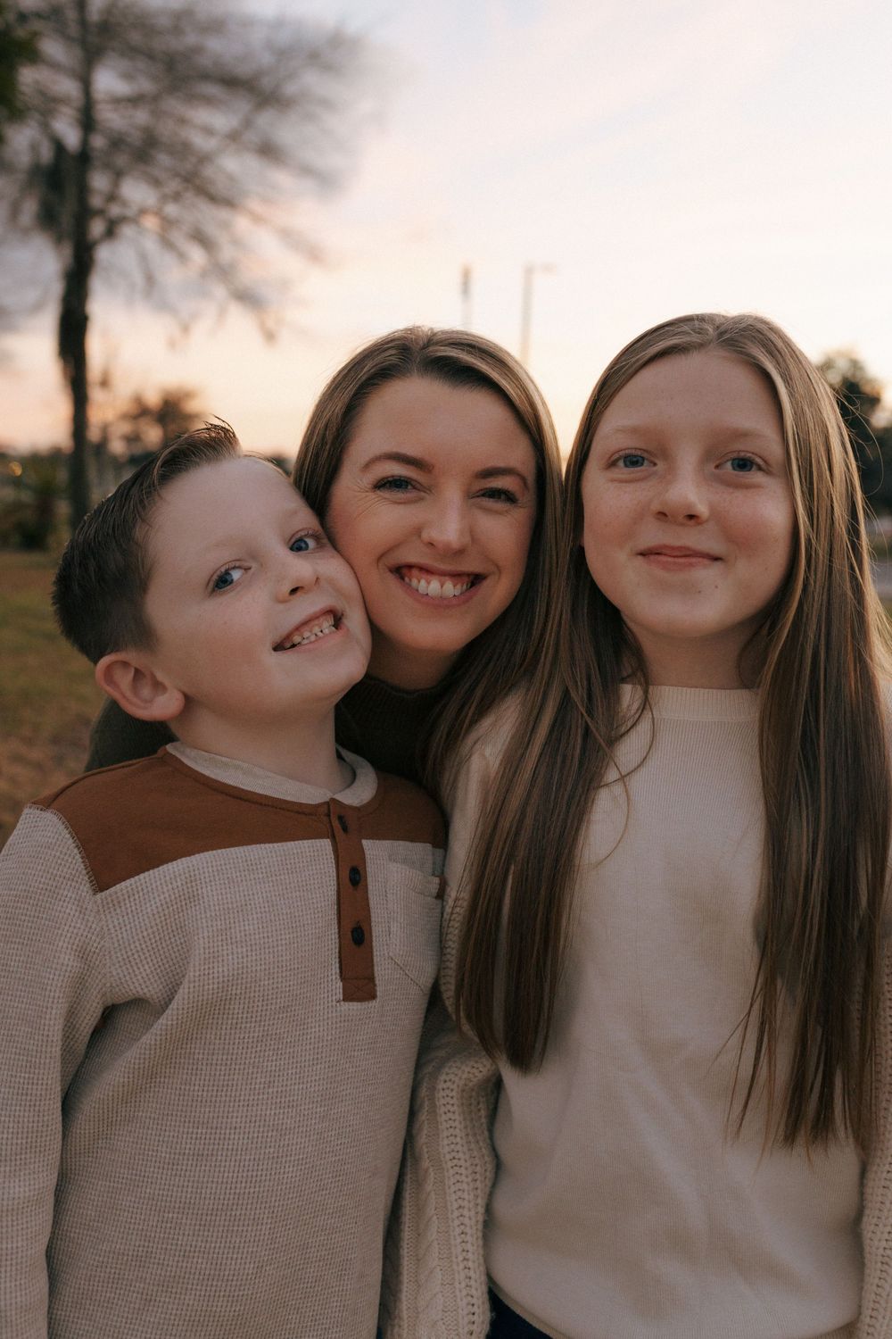 A warm sunset portrait of a family of three with glowing smiles and soft neutral-colored sweaters.