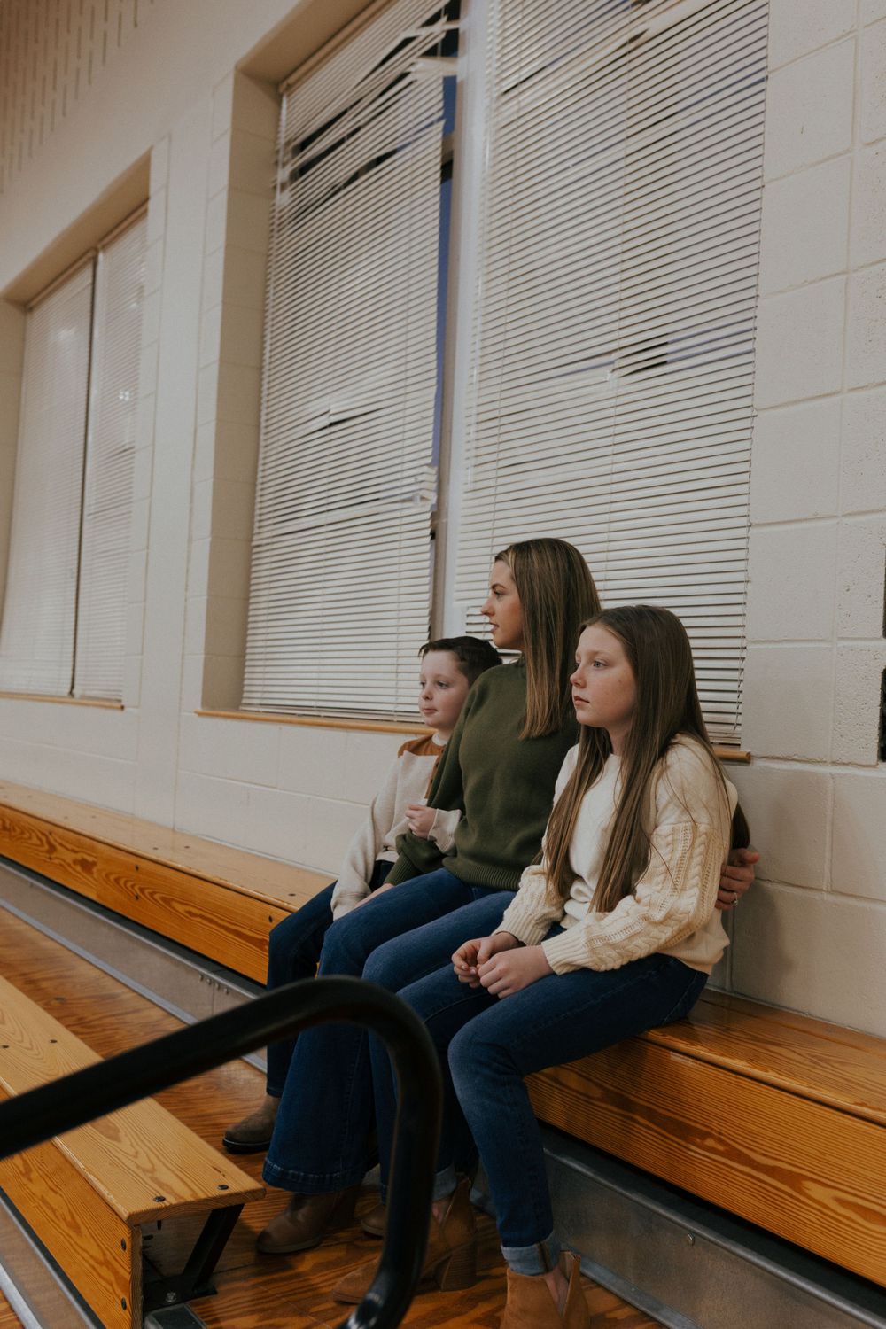 Three people sit on wooden gymnasium bleachers against a white wall with closed blinds in the window.