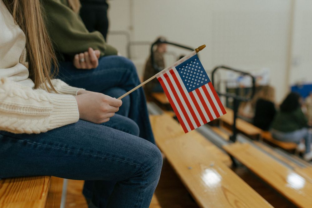 Someone in jeans and a cream sweater holds a small American flag while seated on wooden bleachers.