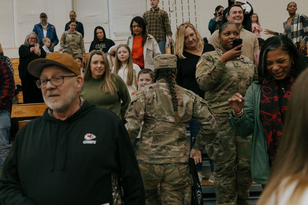A crowd of military personnel and civilians gather in an indoor venue with an American flag visible in the background.